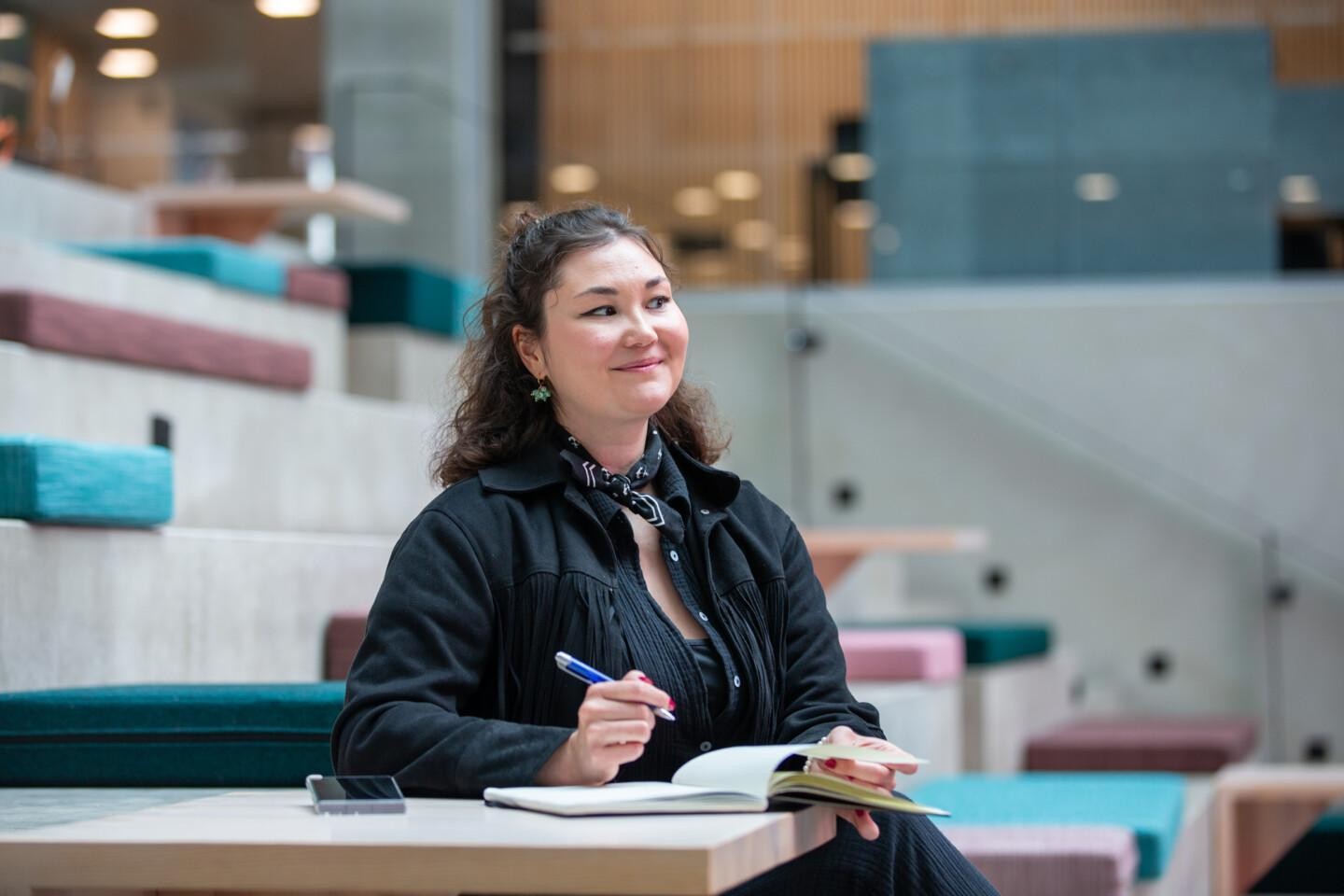 Emma Sarsila istuu pöydän ääressä kynä kädessään ja muistivihko edessään. Hän katsoo hymyillen vasemmalta oikealle.