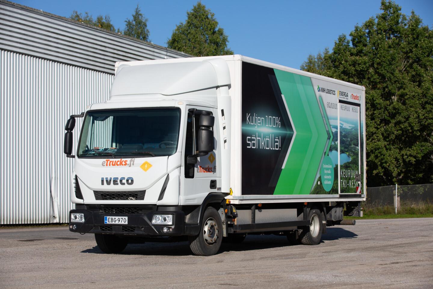 Valkoinen kuorma-auto seisoo asfaltilla teollisuusrakennuksen edessä, kyljessä eTrucks.fi-teksti ja musta-vihreä "Kuljen 100% sähköllä" -teippaus.