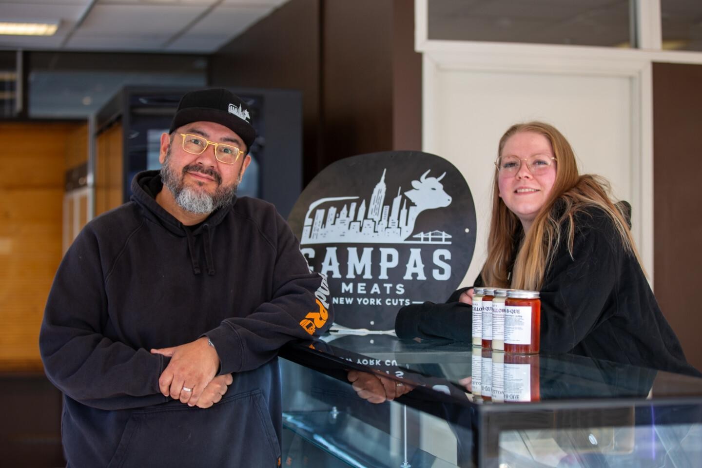 Roberto ja Eveliina Campas seisovat lasivitriinin molemmin puolin hymyillen, taustallaan pyöreä Campas Meats -logokyltti, jossa New Yorkin siluetti ja nauta.