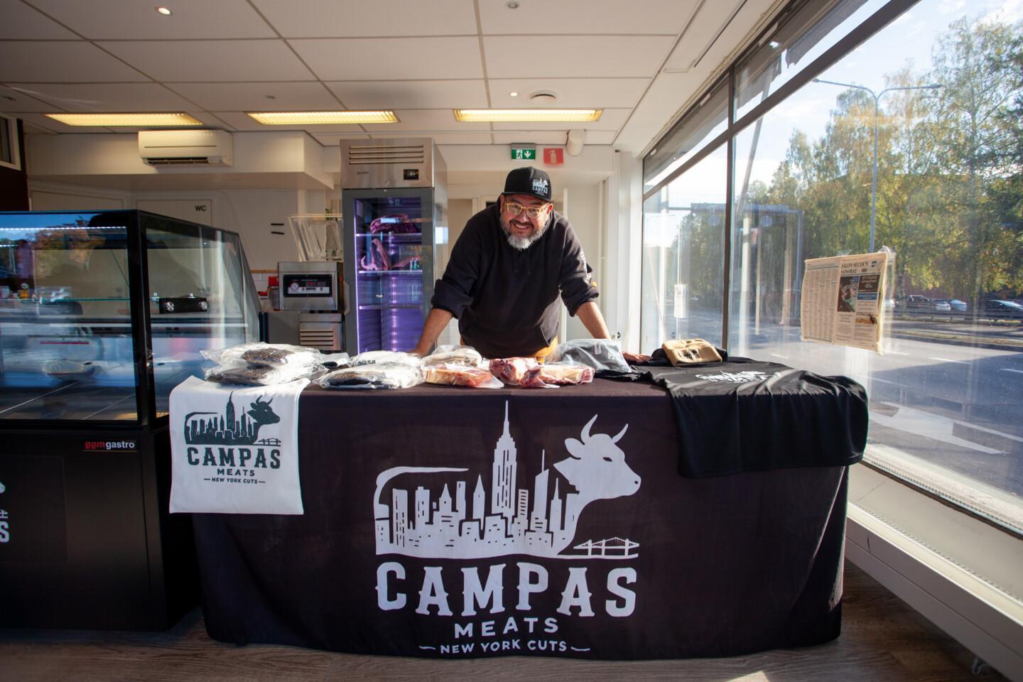 Roberto Campas nojaa hymyillen pöytään, jonka liinassa on Campas Meatsin logo: New Yorkin siluetti ja nauta. Pöydällä on pakattuja lihatuotteita, t-paitoja ja muita tuotteita. Taustalla kylmävitriini ja kaappipakastin, jossa näkyy lihaa, sekä suuret ikkunat, joista avautuu näkymä kadulle.